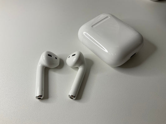 Photo of an AirPods 2nd generation set. Everything is separate and on the white desk: the left AirPod, the right AirPod, and the charging case.
